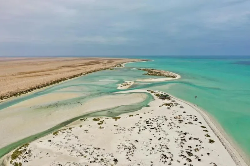 Aerial view of a coastal area with turquoise waters, white sandy beaches, and a narrow waterway winding through the land, with some buildings near the shoreline and sparse vegetation.