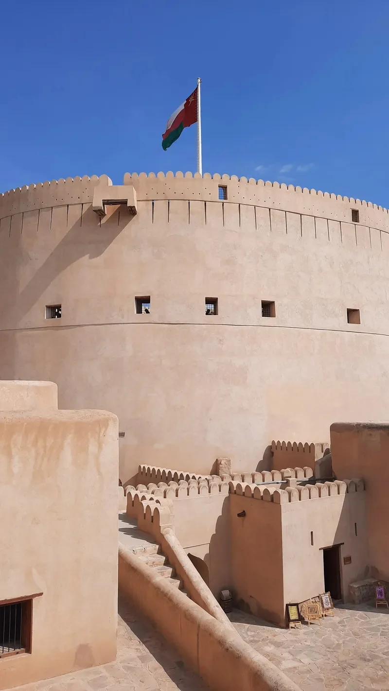 A beige fortress with small square windows and battlements, featuring a flagpole with a red, green, and white flag, under a clear blue sky.