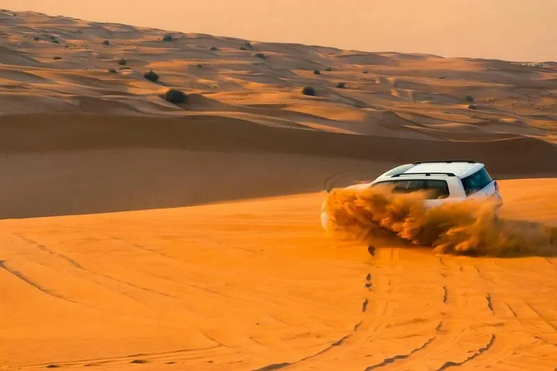 A white SUV driving across a sandy desert, kicking up dust behind it during what appears to be sunset or sunrise, with rolling sand dunes in the background.
