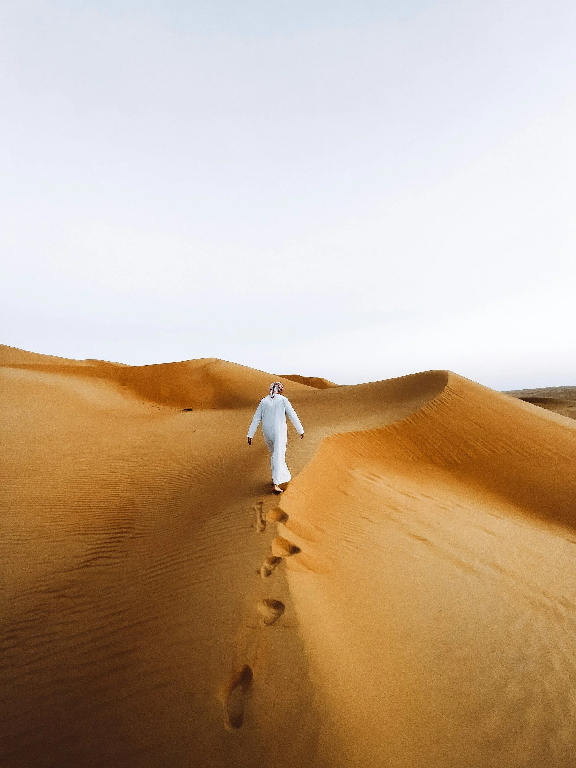 A person dressed in traditional Middle Eastern attire walking across sand dunes in a desert, leaving footprints behind.