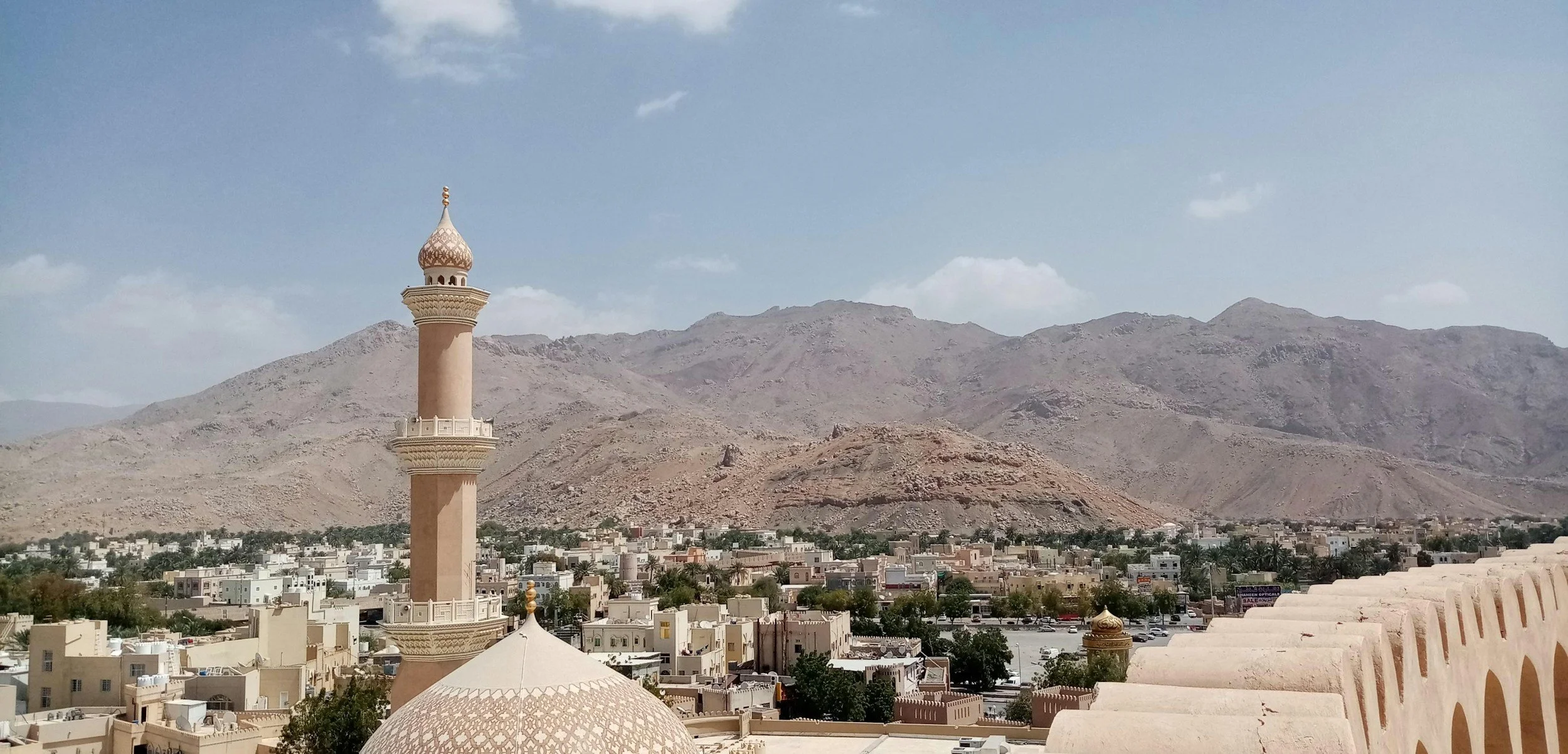 A cityscape with a tall minaret and traditional Middle Eastern architecture in front of rocky desert mountains, under a partly cloudy sky.