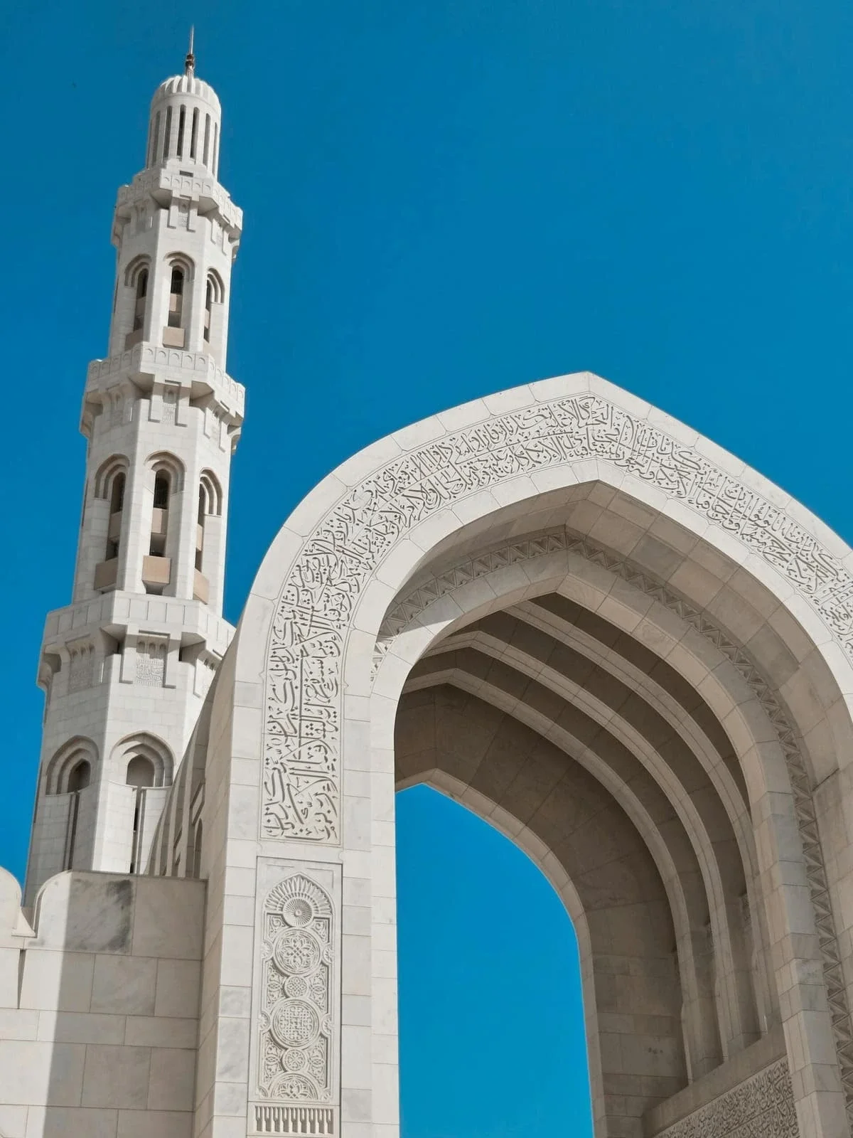 White marble mosque entrance with a tall minaret against a clear blue sky, featuring intricate Arabic calligraphy and geometric patterns.