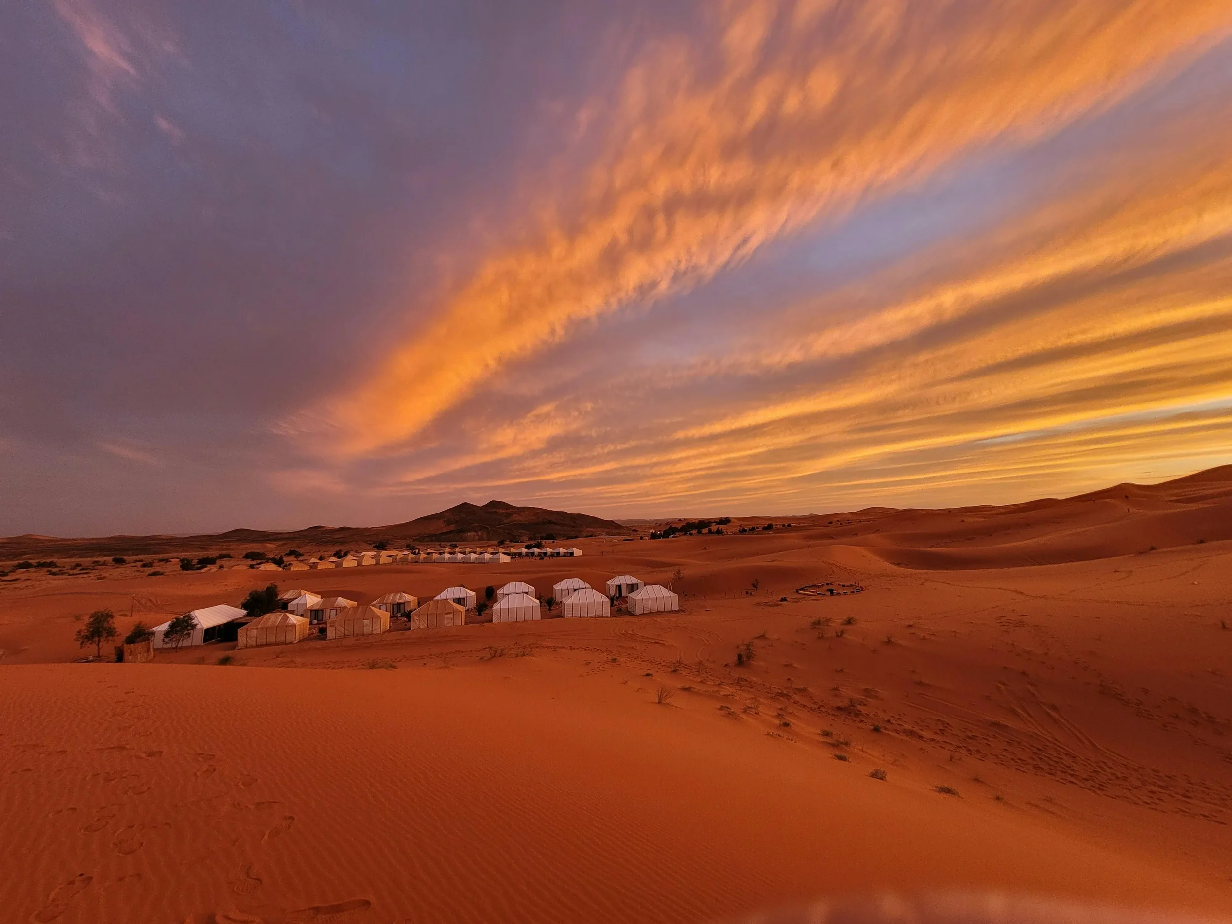 Sunset over a desert landscape with sand dunes, small vegetation, tents, and hills in the background.