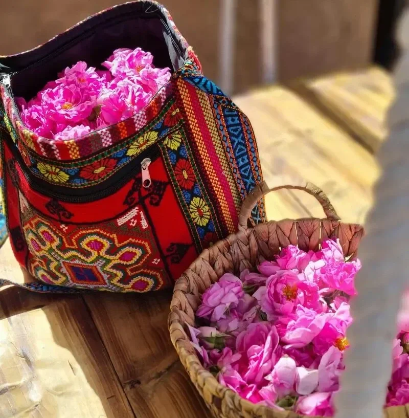 A colorful embroidered bag filled with pink flowers next to a woven basket of pink flowers on a wooden table.