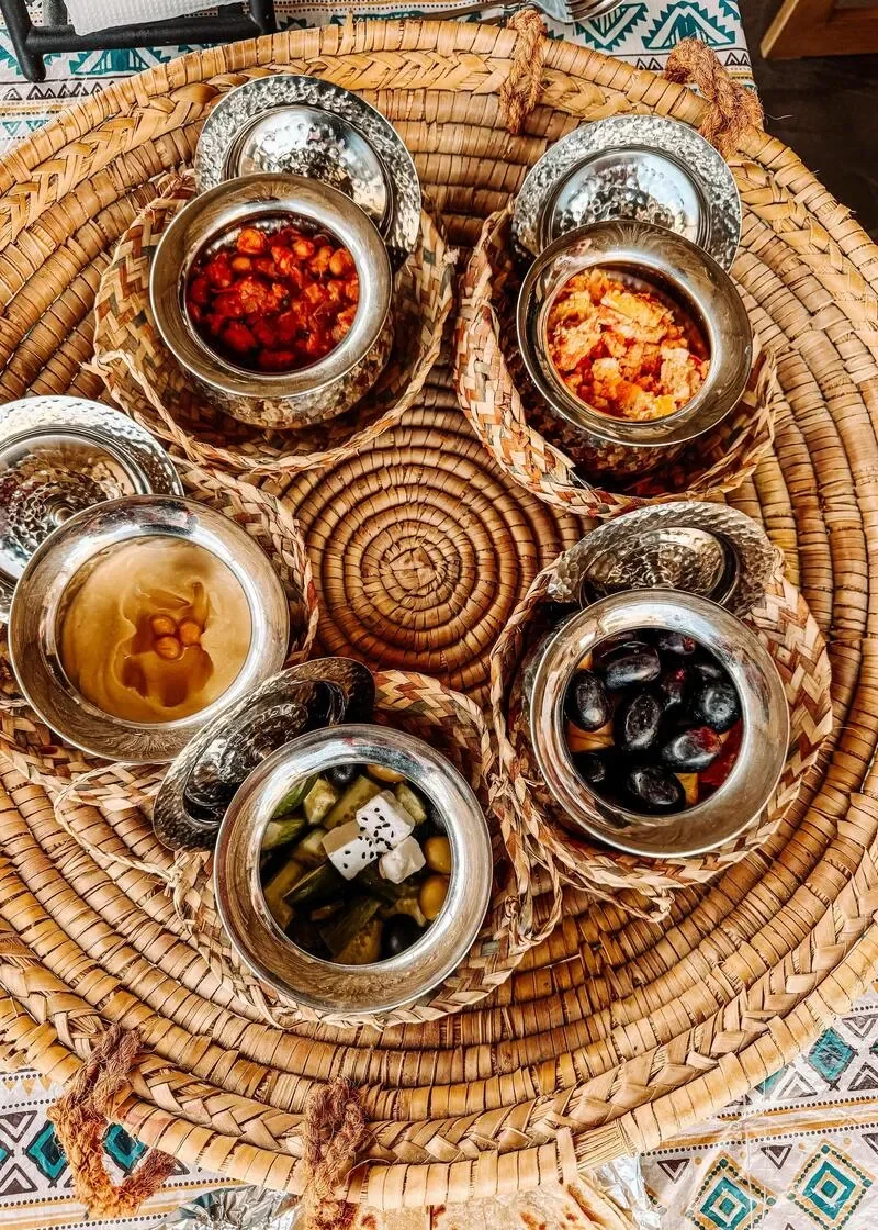 Various types of pickled vegetables and snacks in metal bowls on a woven basket. There are pickled cucumbers, olives, mixed vegetables, fruit slices, and possibly spicy or savory snacks, all displayed on a colorful patterned tablecloth.