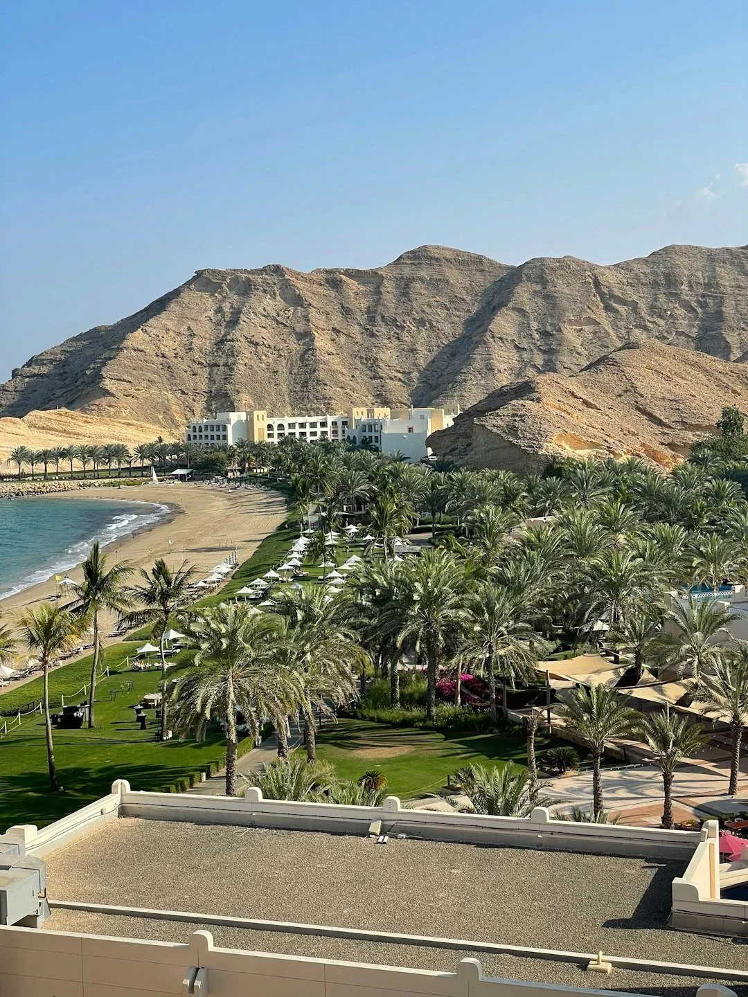 Beachfront resort with palm trees, umbrellas, sandy beach, mountains in the background, and a building complex.