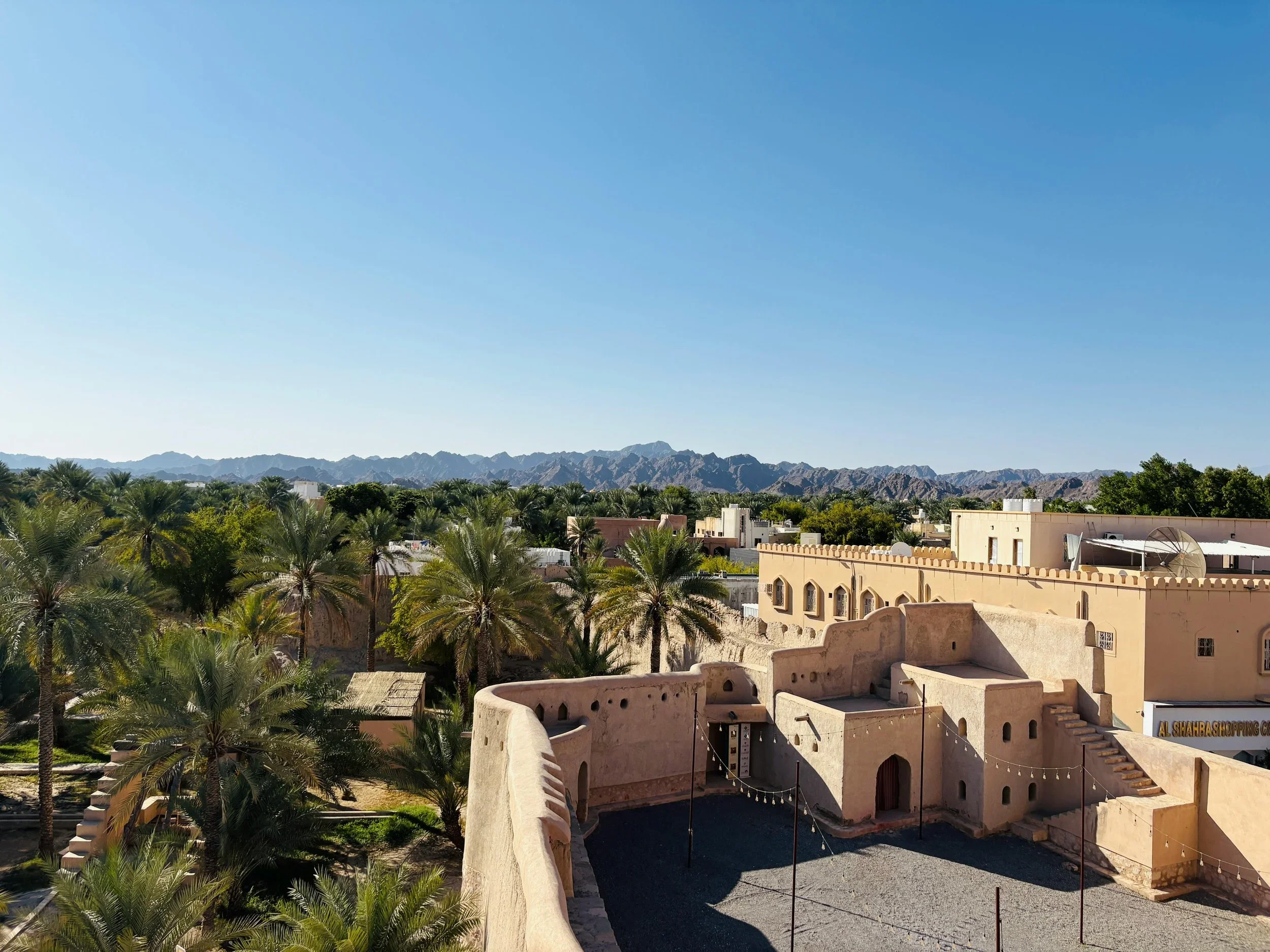 A desert village with traditional adobe buildings, lush palm trees, and mountains in the background under a clear blue sky.
