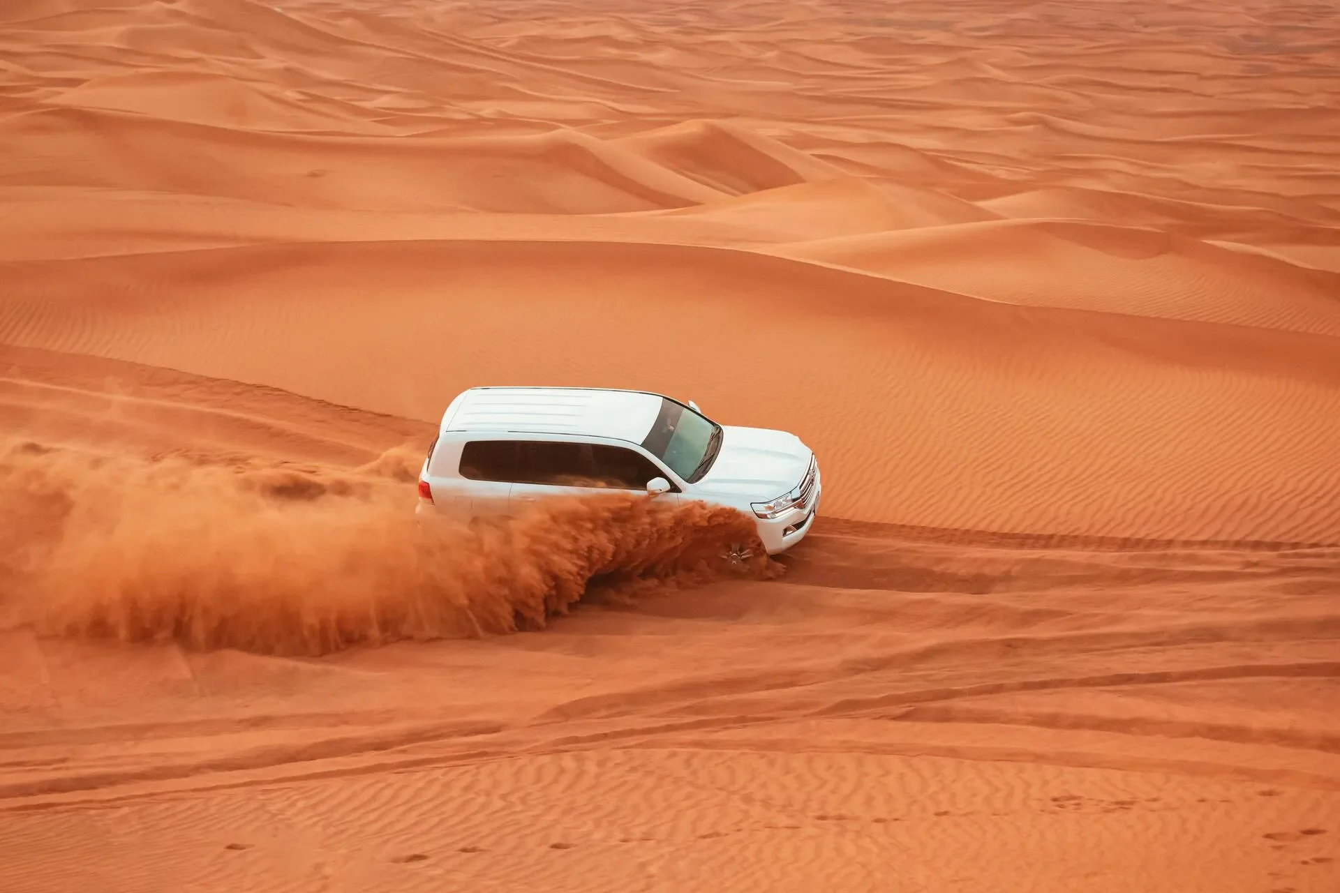A white Toyota SUV driving across desert sand dunes, kicking up dust with a person inside.