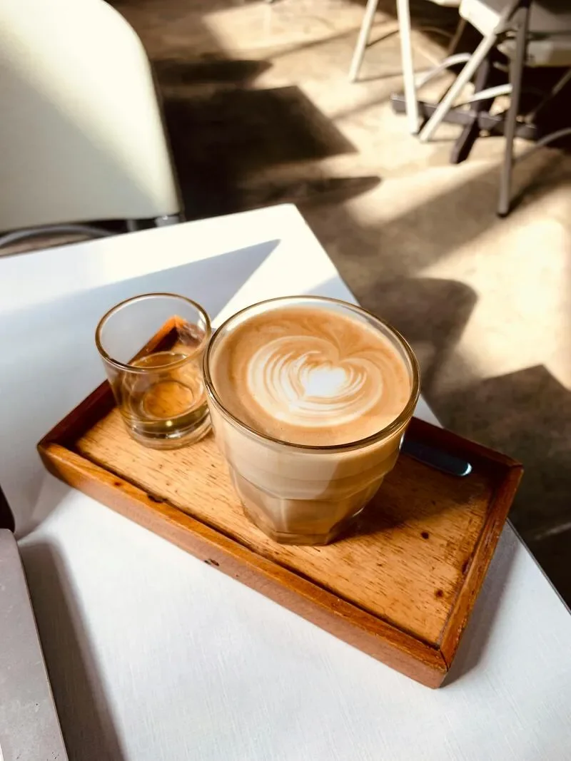 A wooden tray with a glass of latte art and a small glass of water on a white table in a cafe. Shadows and chairs are visible in the background.
