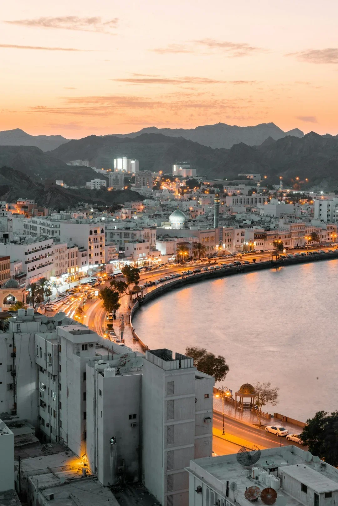 A cityscape at dusk with white buildings along a curved waterway. The sky is pink and orange, with mountain ranges in the background. Streetlights illuminate the streets and building lights glow in the city.