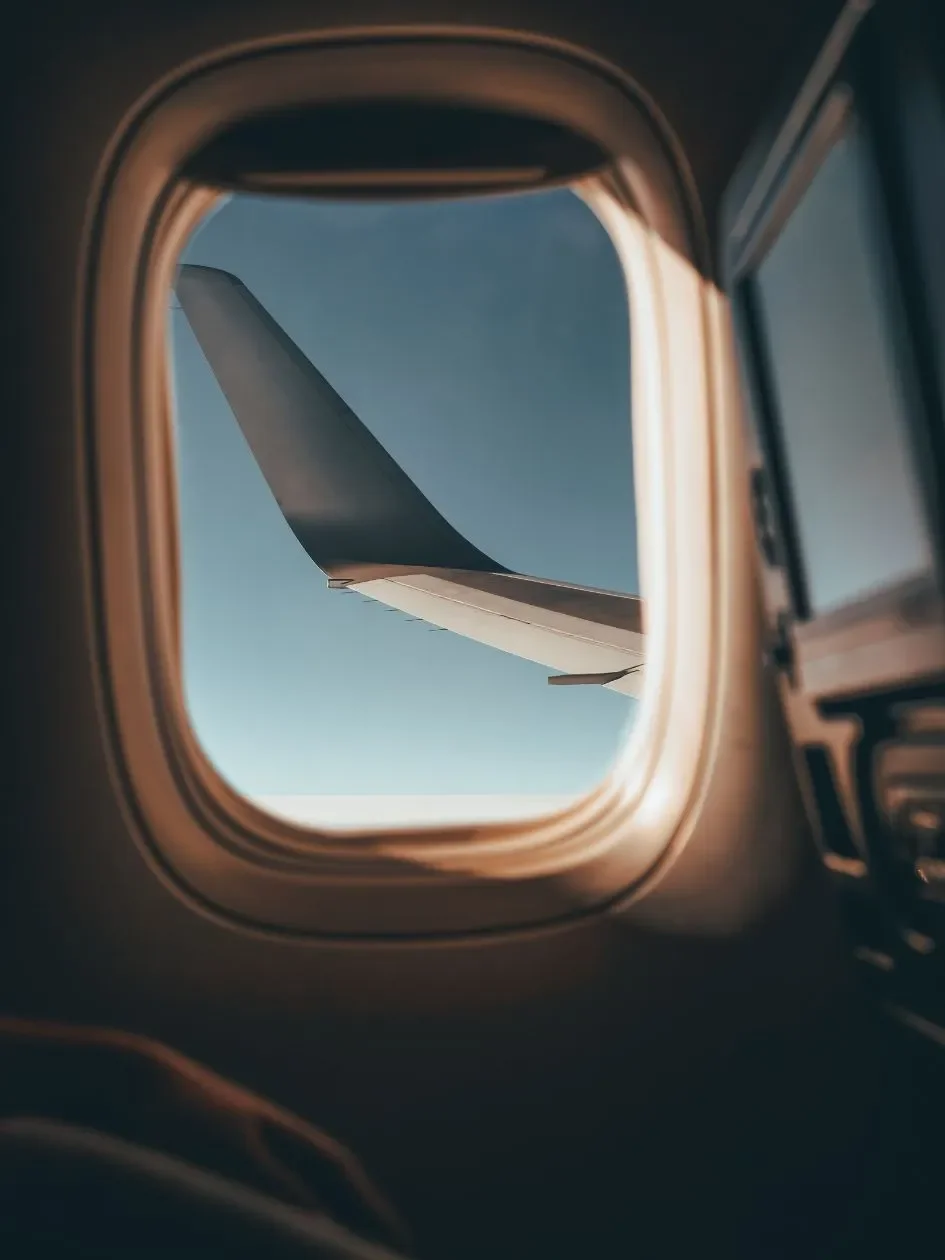 View of an airplane wing through an airplane window during sunset or sunrise, with clouds below and a soft gradient sky.