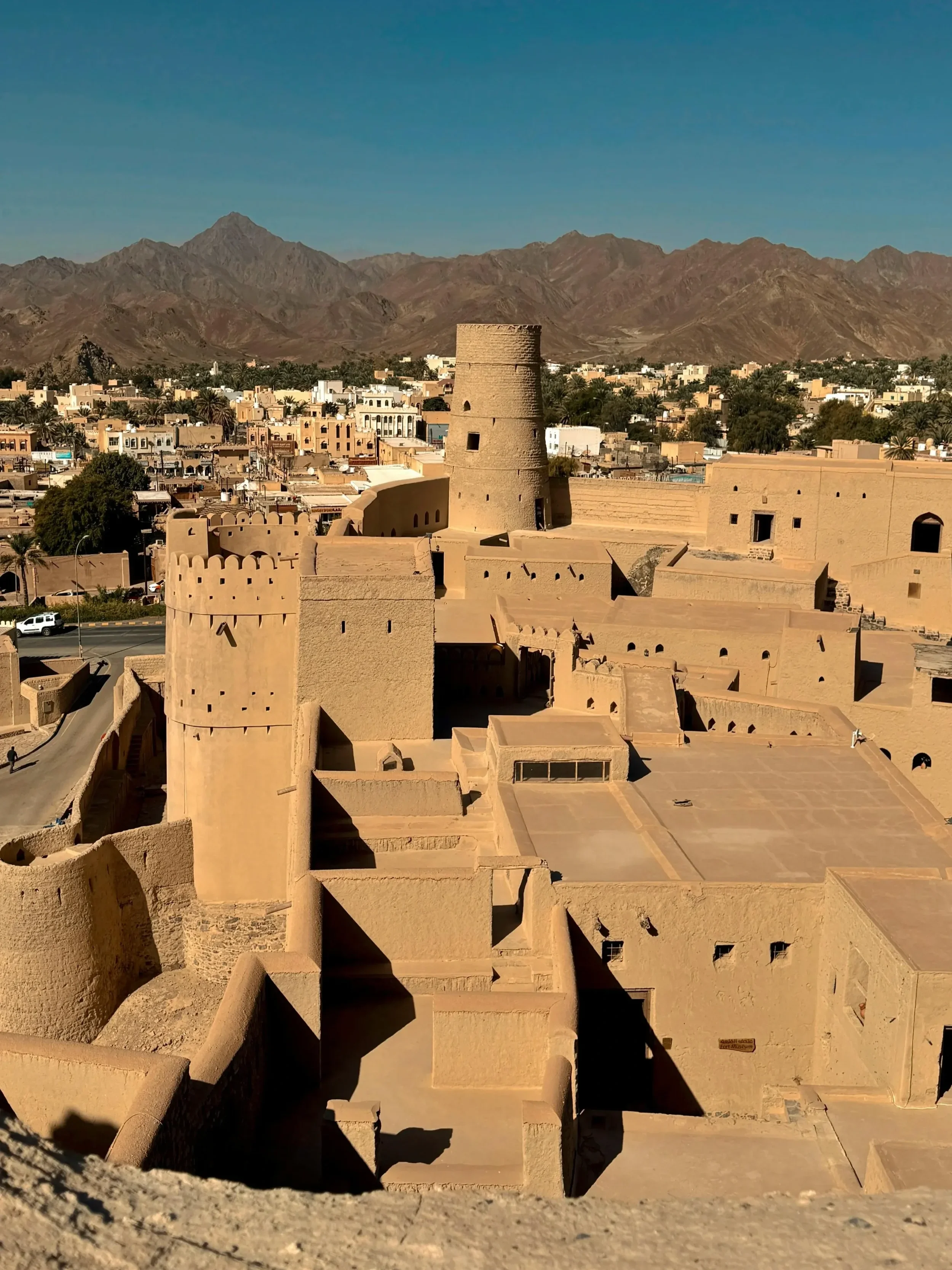 Ancient desert fortress with beige sandstone walls and towers, nestled in a dry, mountainous landscape under a clear blue sky in a Middle Eastern region.
