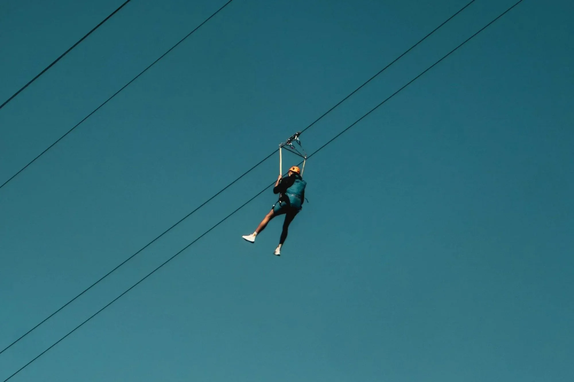 A person is riding a zip line against a clear blue sky, wearing a helmet and safety harness.