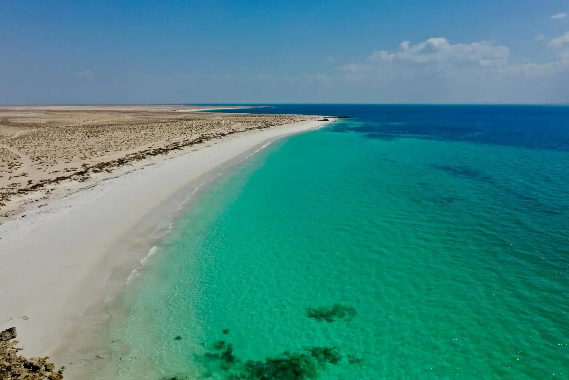 Aerial view of a beach with white sand, turquoise water, and desert terrain in the background under a blue sky with scattered clouds.