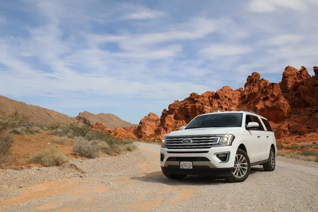 White Ford SUV parked on a dirt road in a desert landscape with red rock formations and a partly cloudy sky.