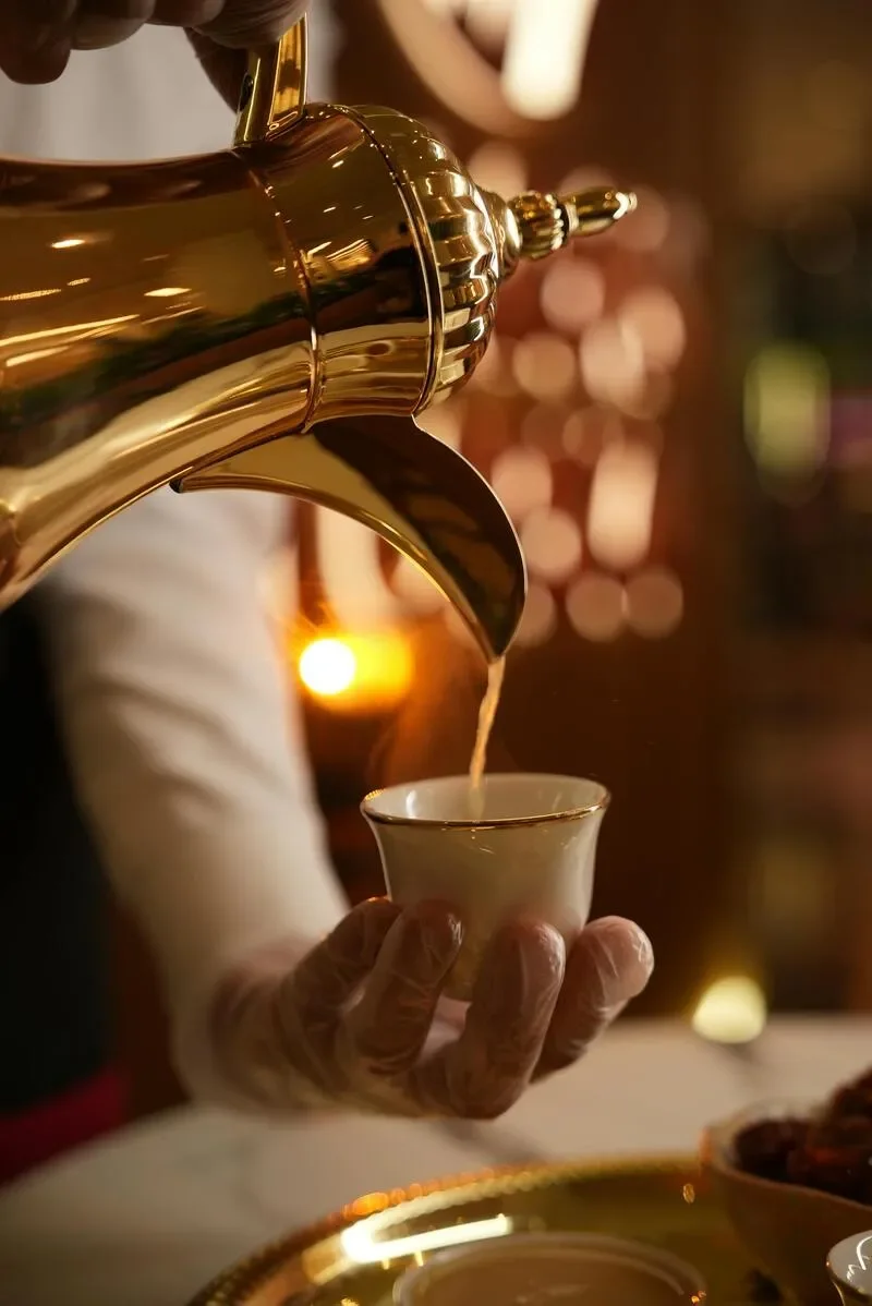 A person holding a white cup while pouring a beverage from a gold teapot. The scene appears warm and elegant, with a blurred background and soft lighting.
