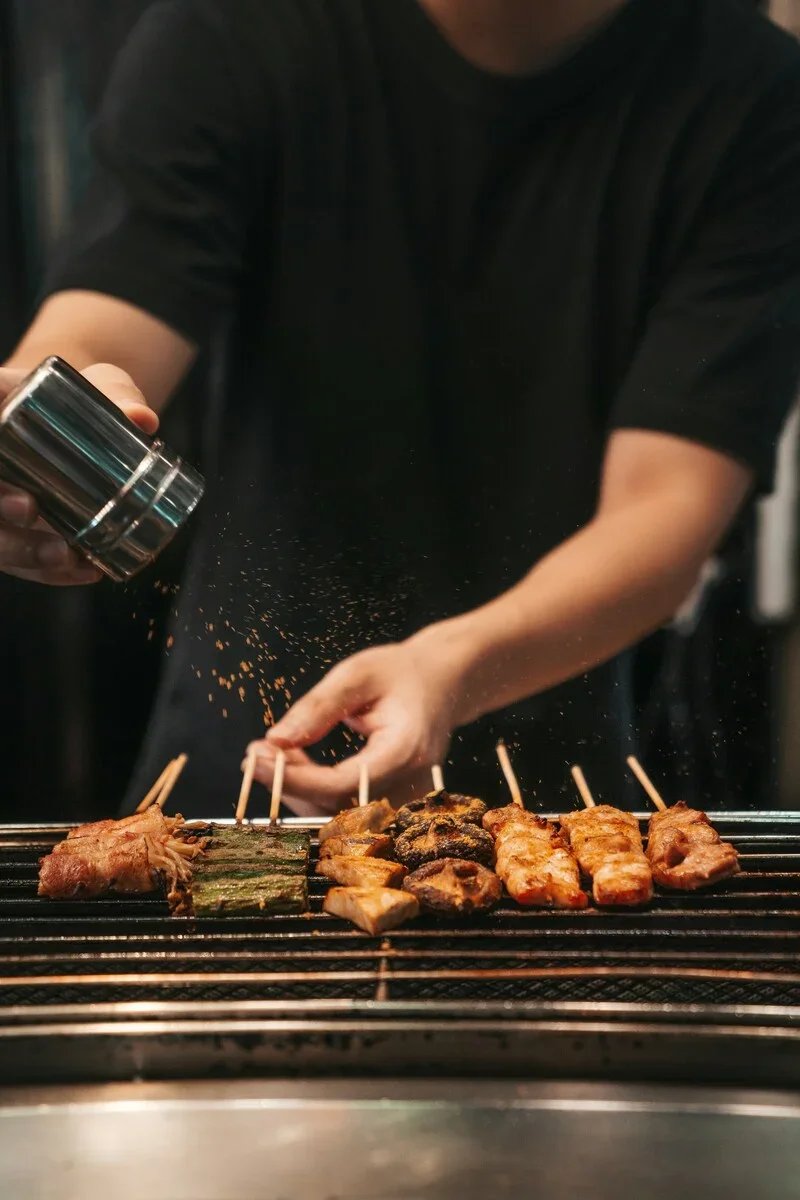 A person wearing a black shirt grilling skewered meat and vegetables on a barbecue grill, sprinkling spices over the food.