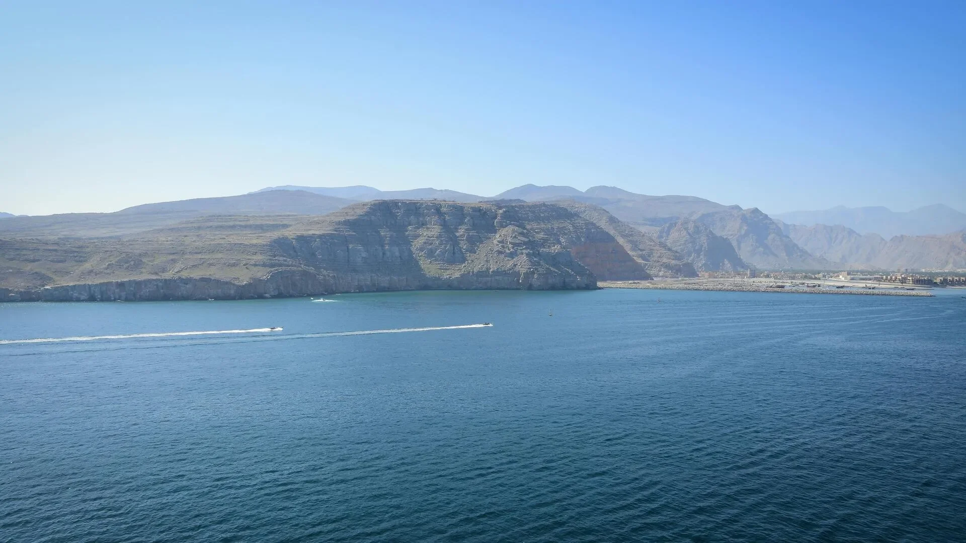 A large body of water with two boats creating wakes, with rocky hills and mountains in the background under a clear blue sky.