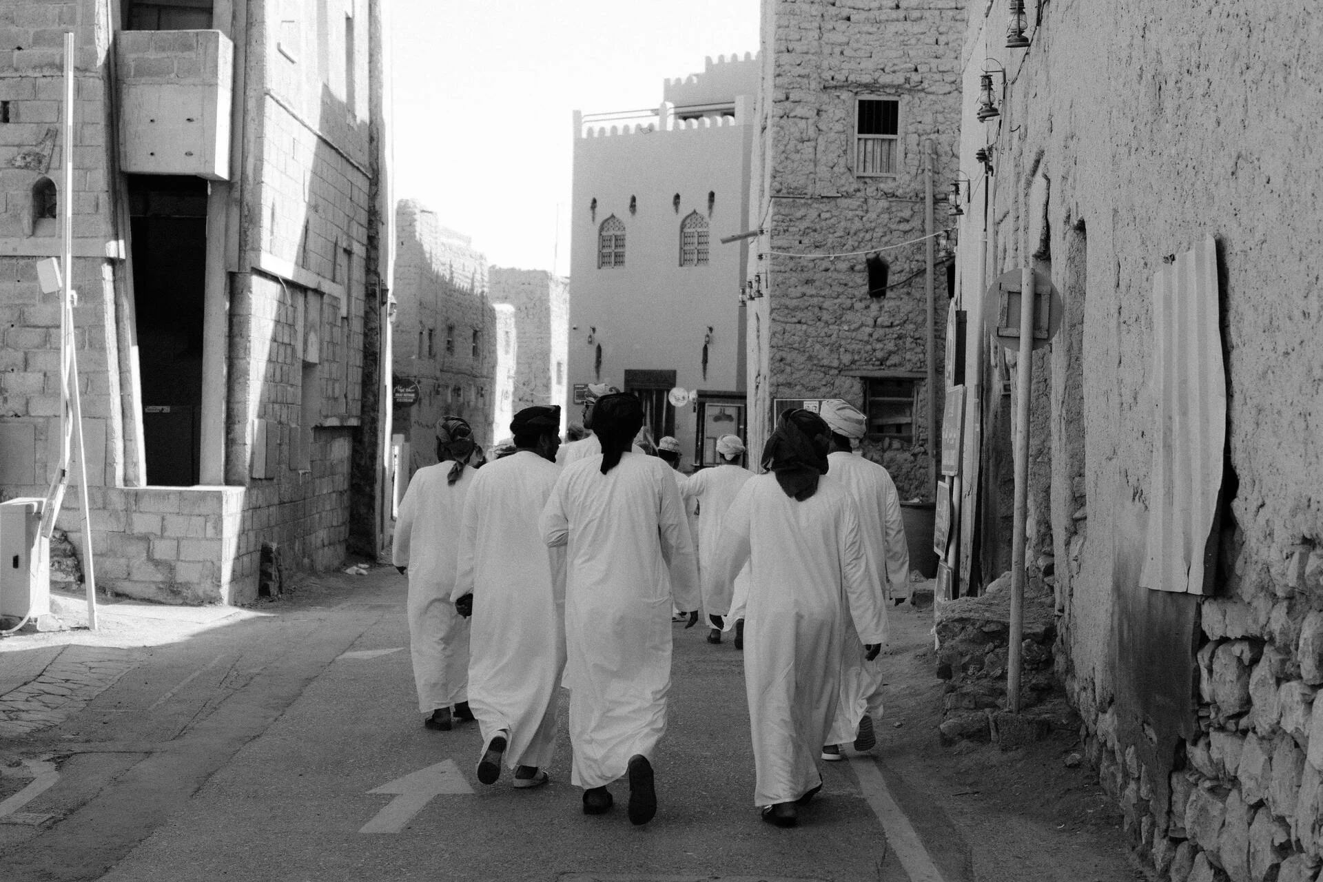 A group of men dressed in traditional white robes walking down a narrow street lined with old buildings, some of which are under construction or renovation, in a historic town.