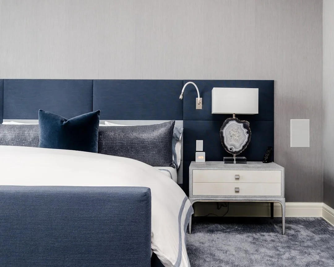Close-up of a modern hotel room with a bed, blue headboard, pillows, a nightstand with a lamp, a clock, a remote, and a textured stone decoration, against a light grey wall.