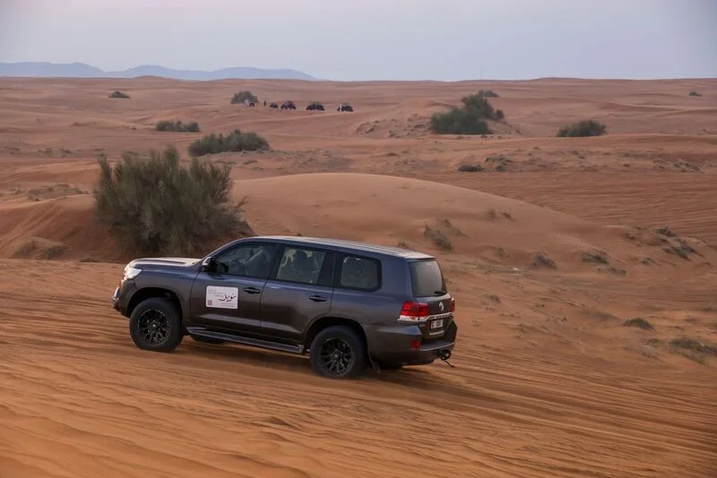 A black SUV driving on sandy desert dunes with sparse vegetation and a distant group of off-road vehicles in the background.