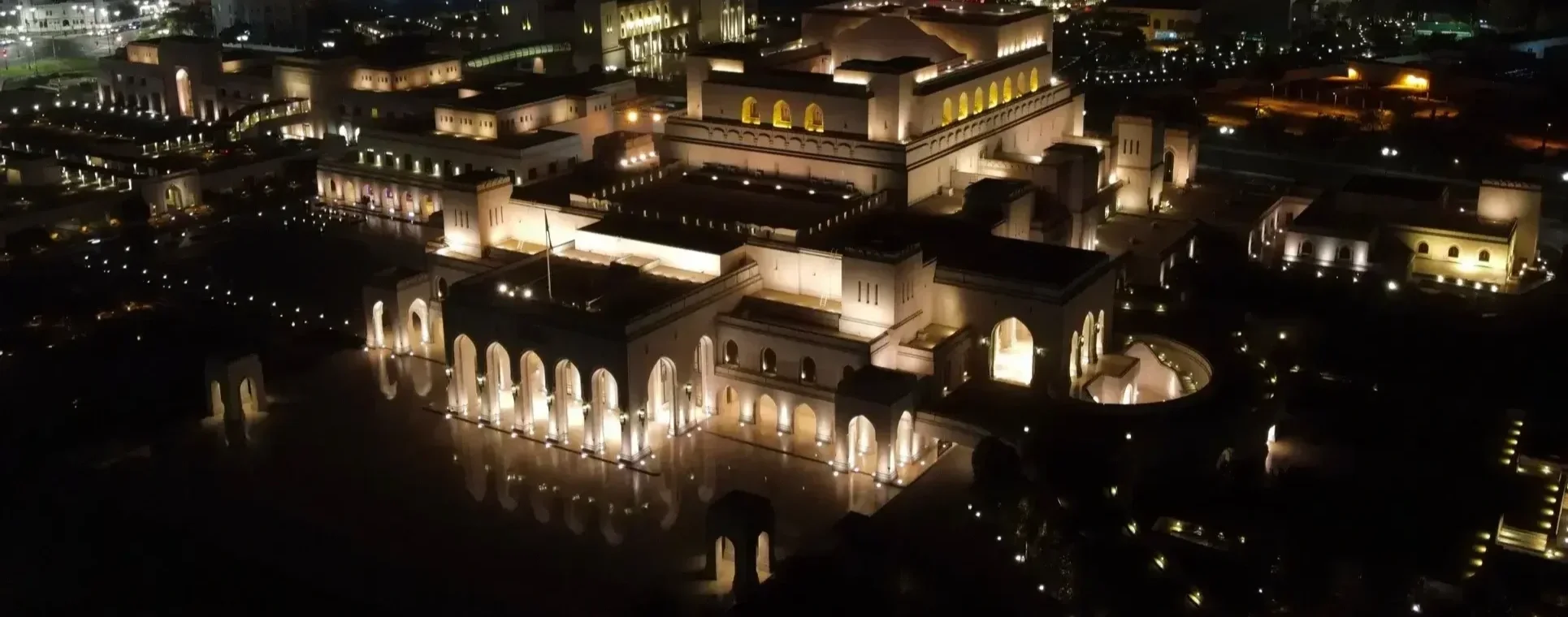 Nighttime aerial view of an illuminated Royal Opera House Muscat with arches and multiple levels.