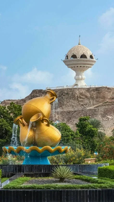 Golden decorative vases pouring water into a fountain with a large white decorative building on a hill in the background, surrounded by greenery.