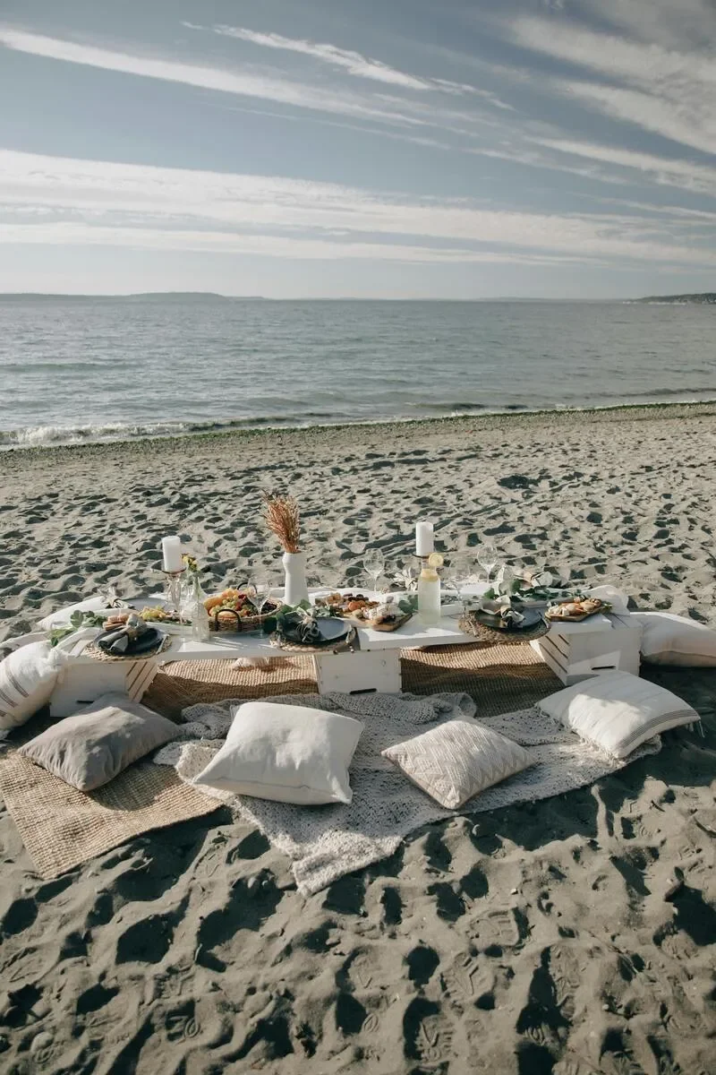 A beach scene with a low table set with food, drinks, and decorations, surrounded by cushions and pillows on the sand, with the ocean and sky in the background.