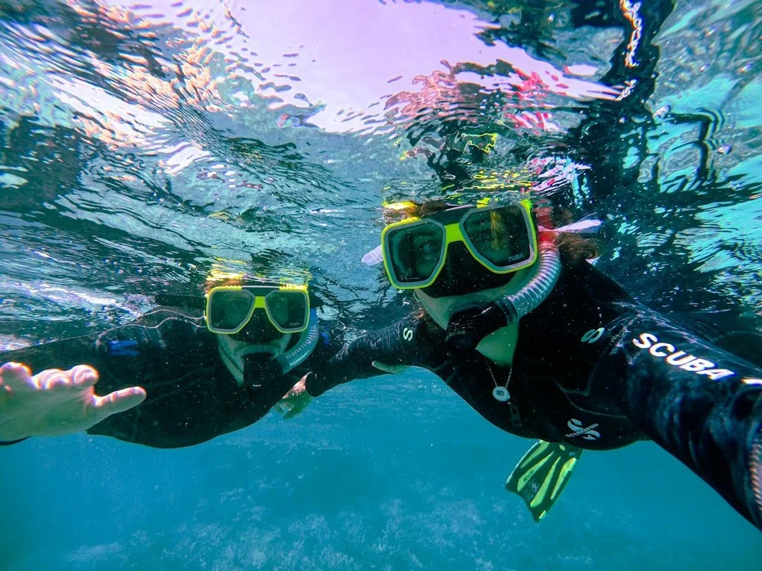 Two snorkelers wearing masks and snorkels swimming underwater, with a colorful underwater scene and ripples on the surface above.