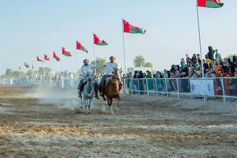Two men in traditional attire riding horses on a dirt track during daytime, with a large crowd of spectators behind a barrier, and multiple flagpoles with flags in the background.