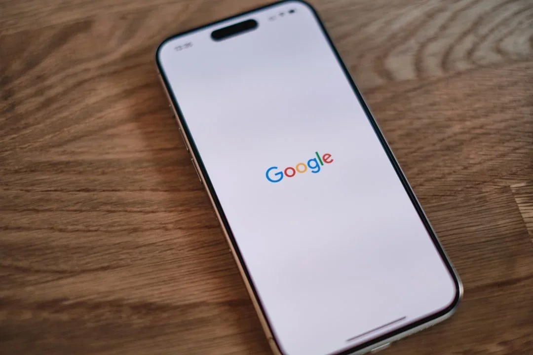 A modern smartphone with a notch at the top of the screen, displaying the Google logo on a white background, on a wooden surface.