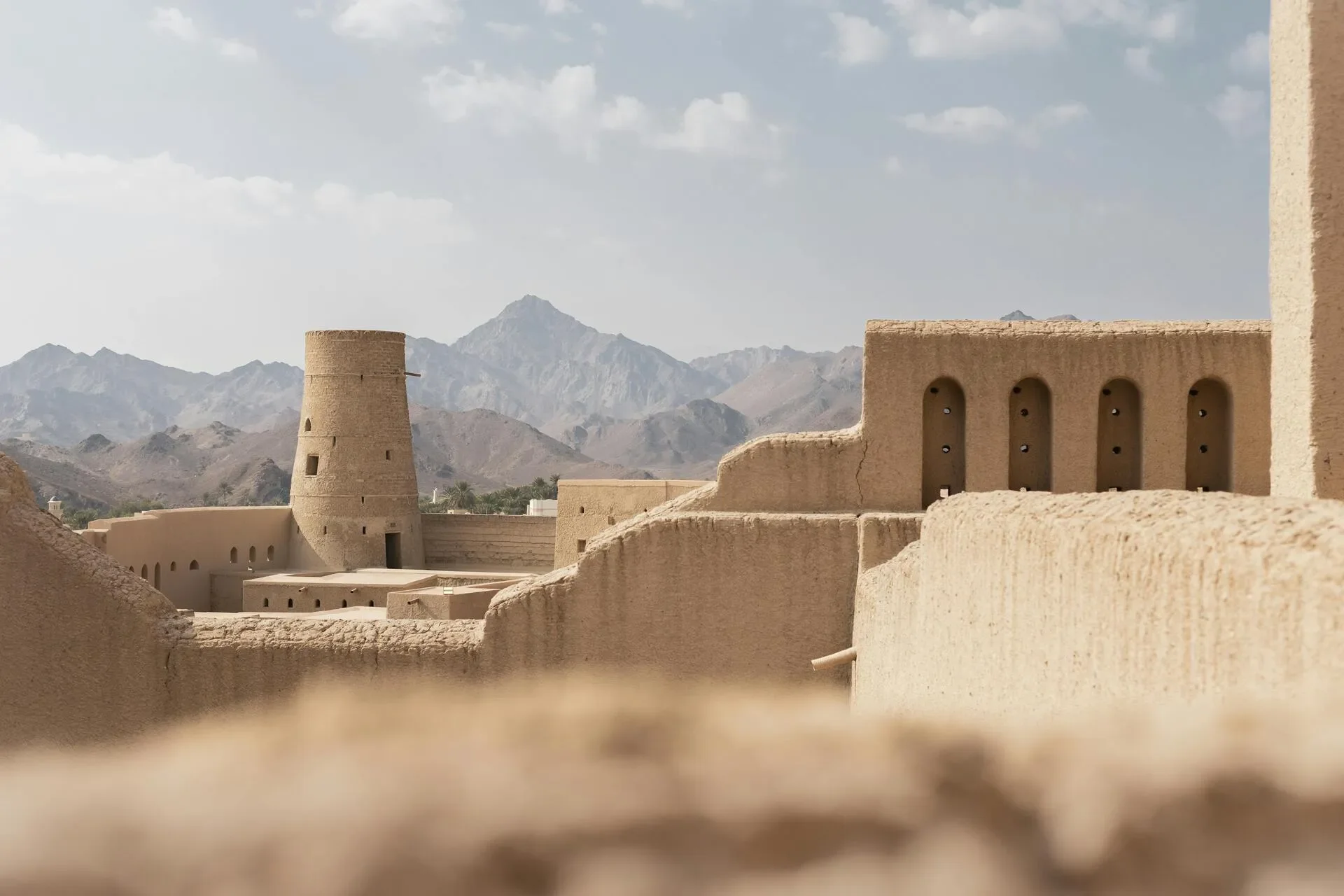 Ancient desert fortress with adobe walls and towers set against mountainous background under cloudy sky.