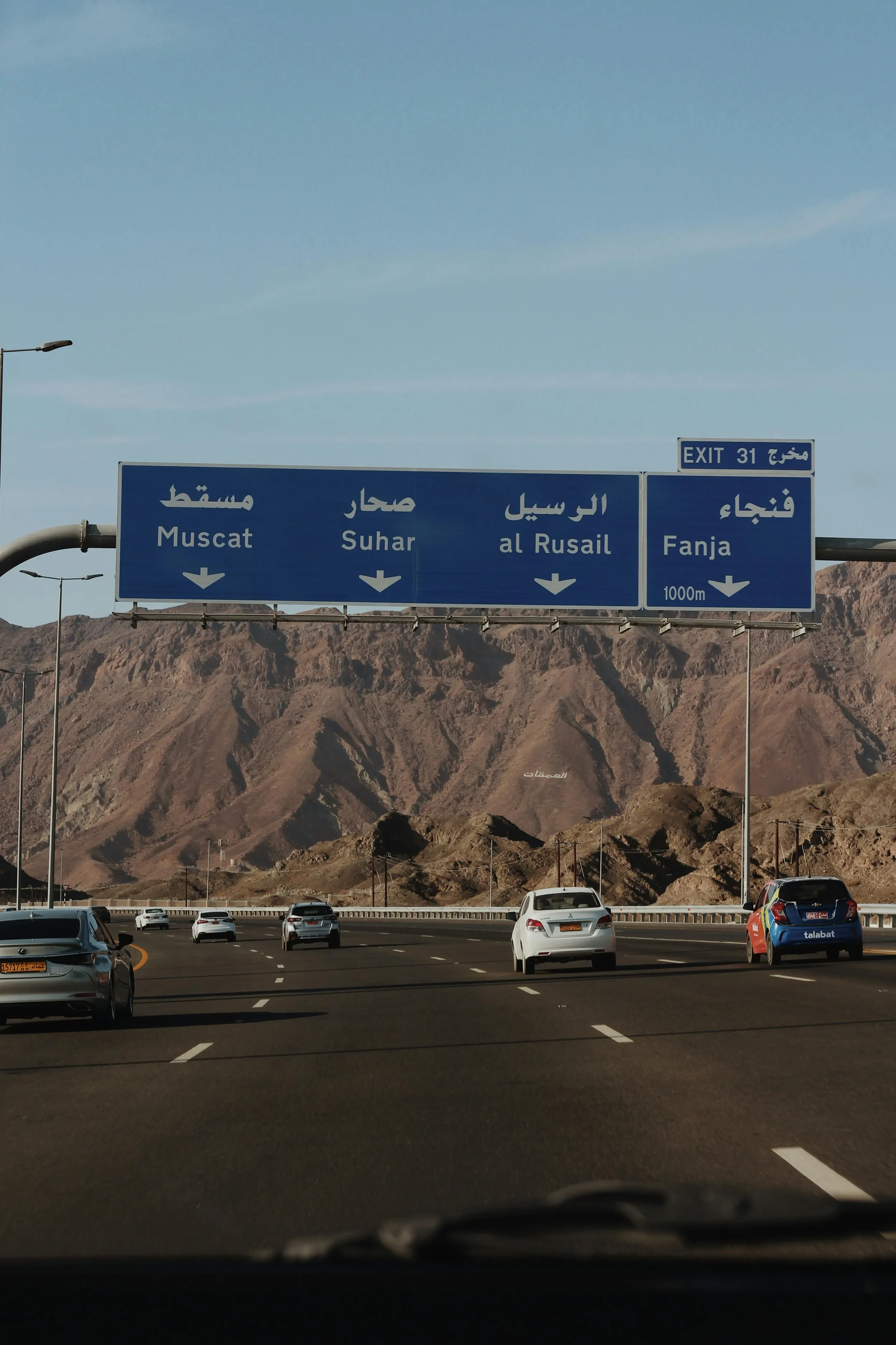 View from inside a car on a highway with parked cars, mountainous desert landscape in the background, and a blue overhead road sign indicating routes to Muscat, Suhar, Al Rusaid, and Fanja.