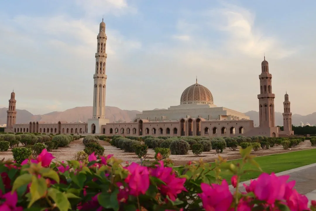 The image shows a mosque with a large central dome and four tall minarets, surrounded by flowering plants and bushes, with mountains in the background and a partly cloudy sky overhead.
