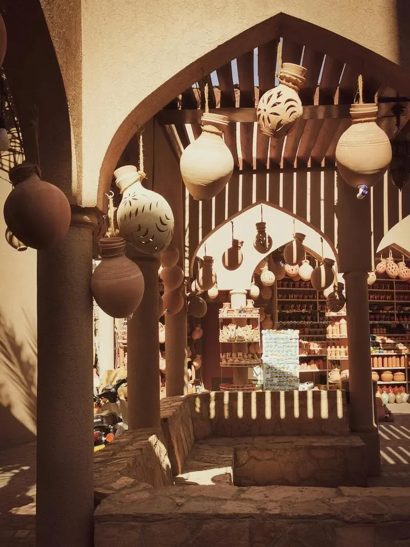 View of a marketplace or shop with hanging ceramic lamps and pottery, arched entrance, shelves filled with ceramic bowls and vases, and warm sunlight.