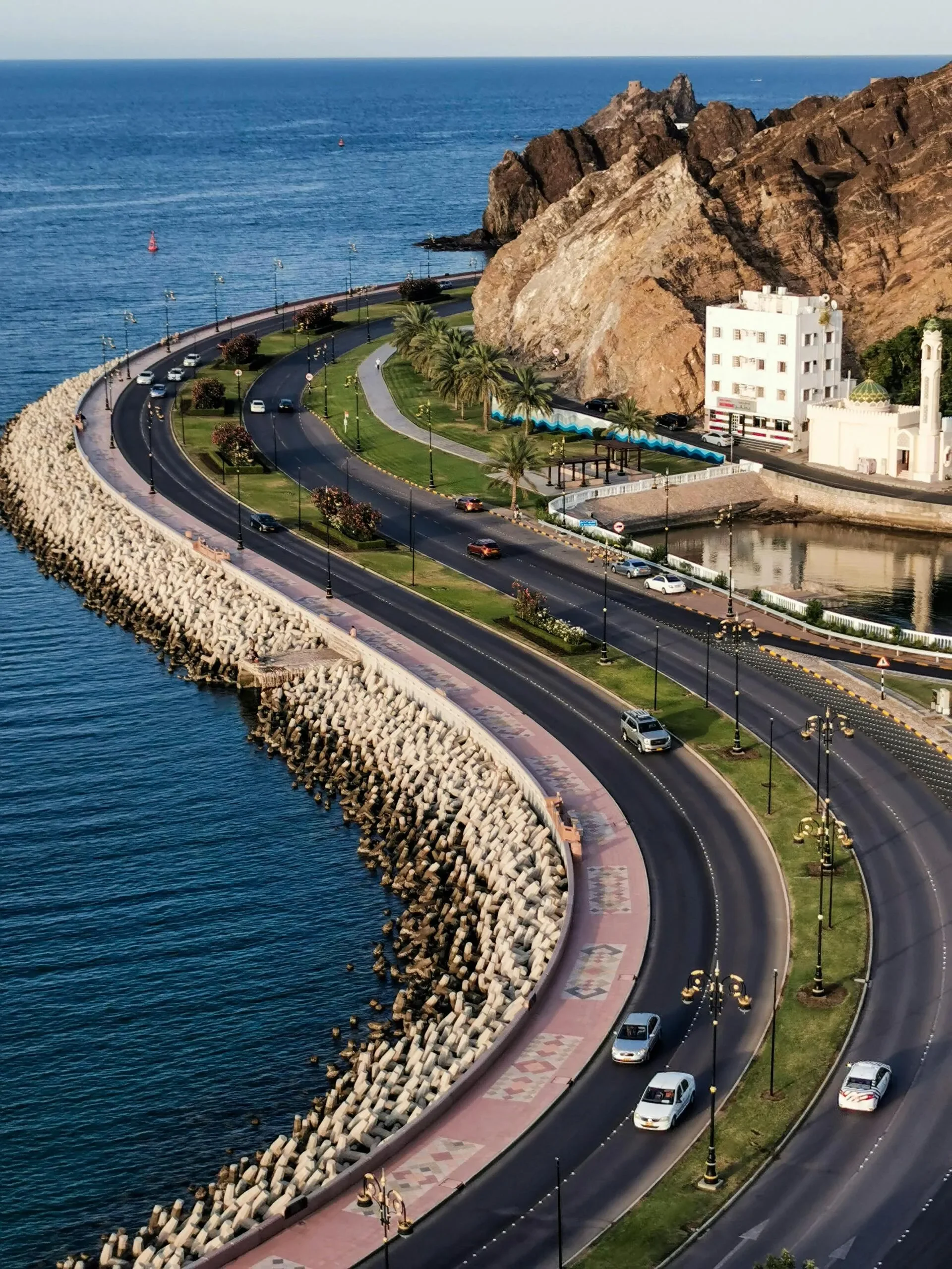 A coastal road with cars, lined with street lamps and palm trees, runs along the edge of the sea, with rocky cliffs in the background and a small building with a minaret nearby.