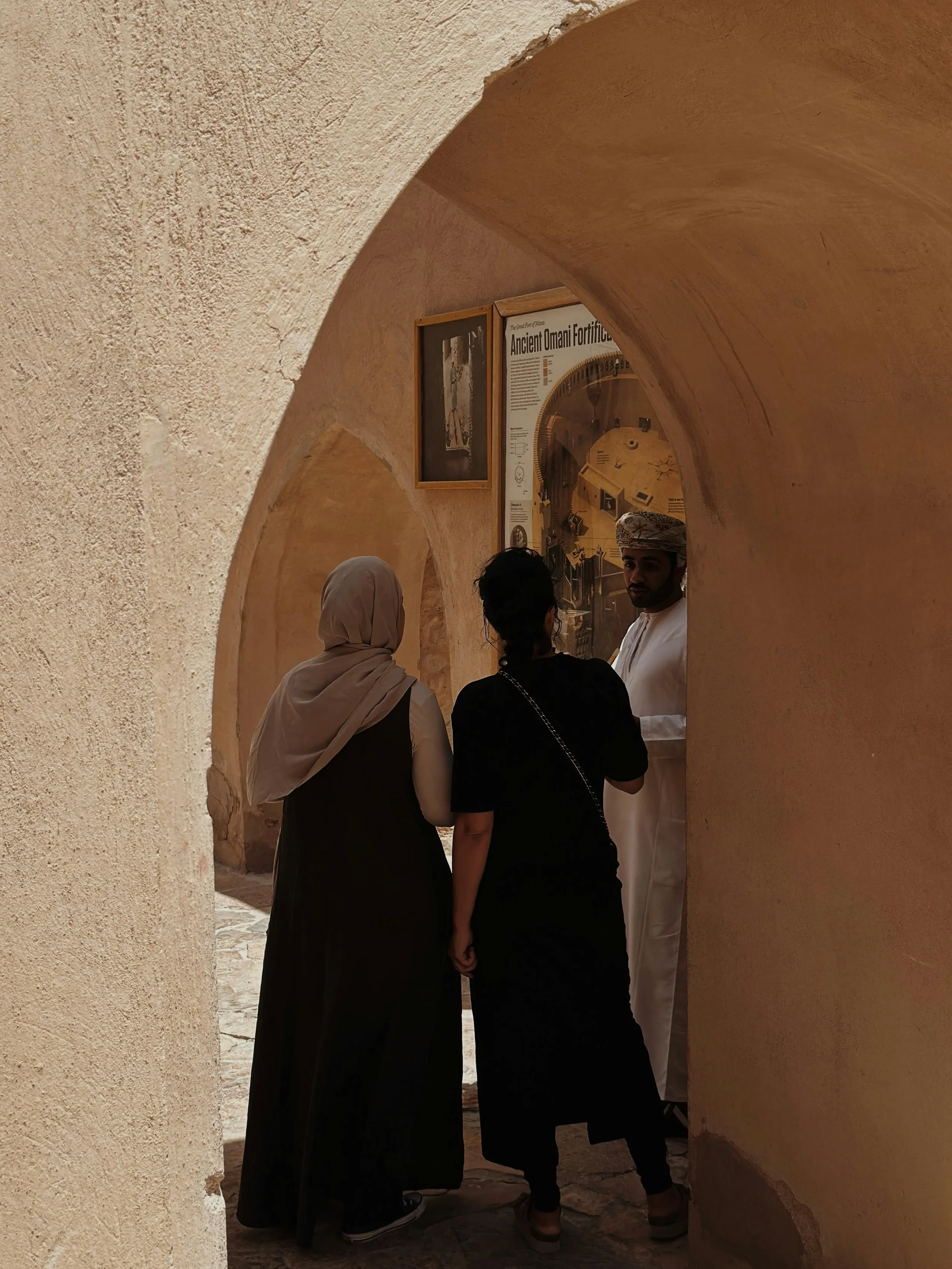 Three people standing and talking inside a narrow hallway or archway with beige textured walls. One person is wearing a traditional turban, another has a headscarf, and the third has short hair. There are framed informational posters on the wall behind them.