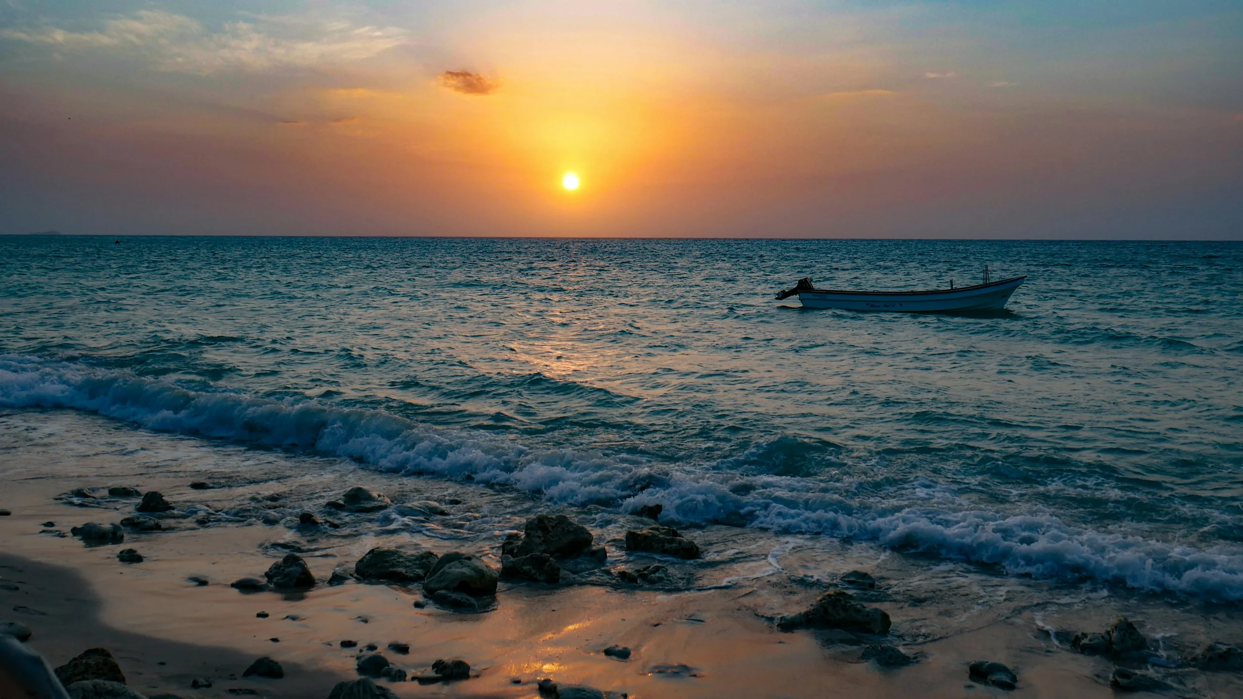 A boat floating on the ocean during sunset with the sky painted in warm colors and gentle waves reaching a rocky shoreline.