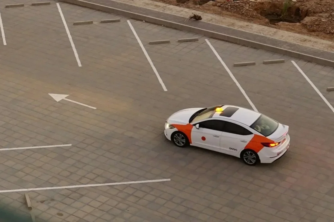 An empty parking lot with a single car, which is a white sedan with orange markings, a yellow roof light, and a red dot on the side.