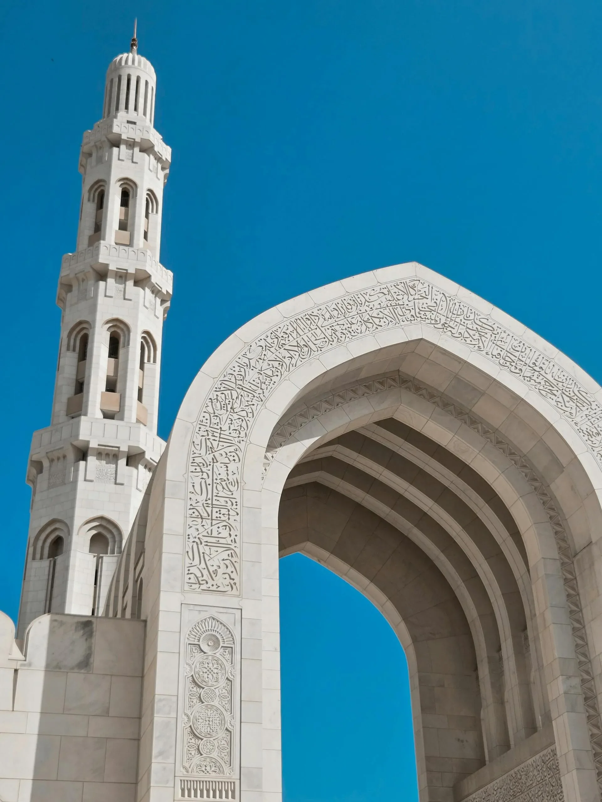 Archway and minaret of a white mosque with intricate Arabic calligraphy, set against a bright blue sky.