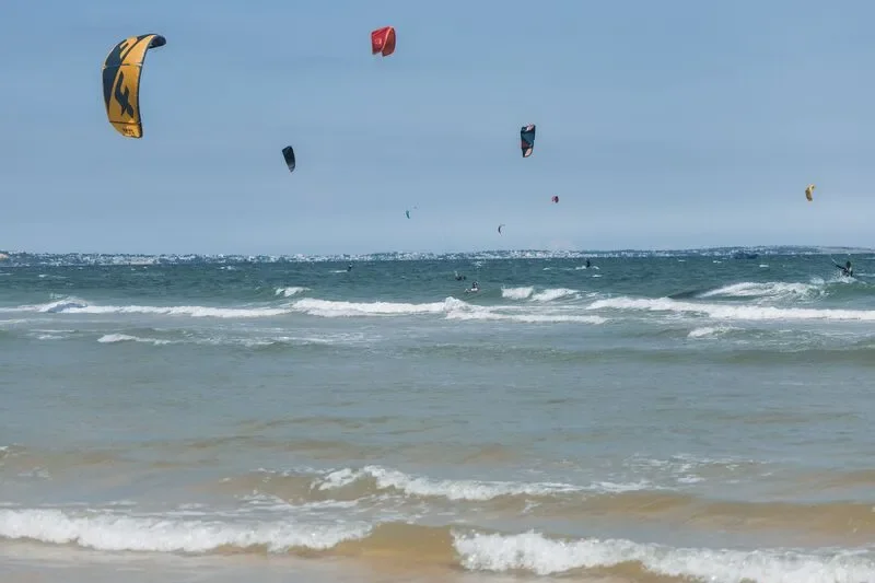 Several kite surfers are riding the waves at the beach, with colorful kites in the sky and a distant shoreline visible on the horizon.