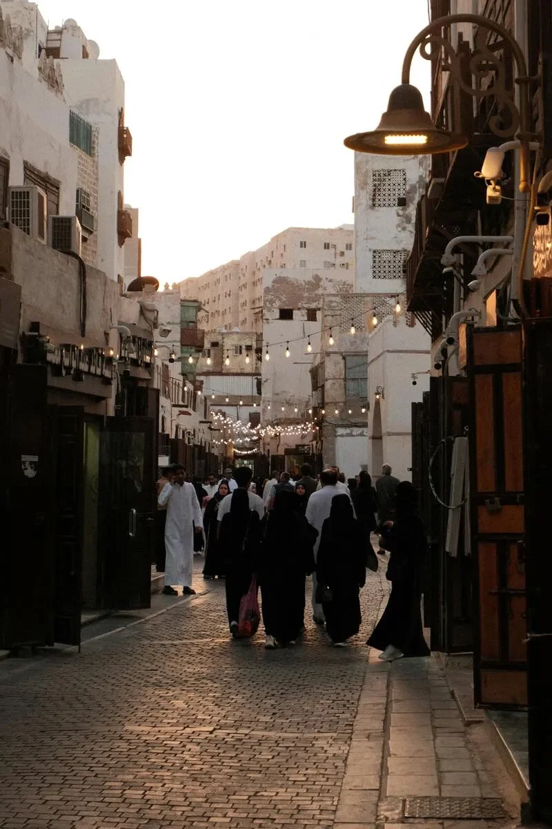 A busy street scene during dusk with pedestrians walking and hanging string lights overhead. The buildings are worn and weathered, with some windows and air conditioning units visible.