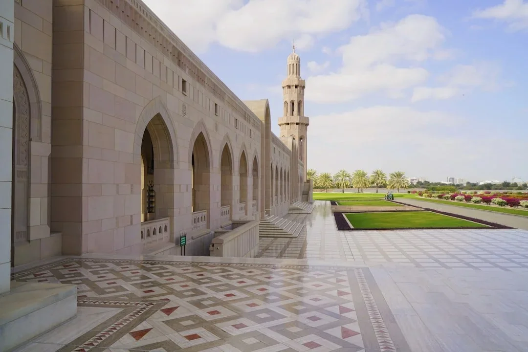View of an Islamic architectural building with arches and a minaret, surrounded by a landscaped garden with pathways, palm trees, and flowers.