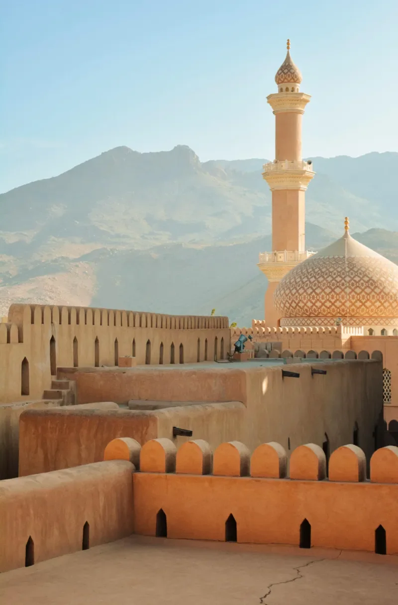 A desert fortress with terracotta walls and a minaret, set against a mountainous background under a clear blue sky.