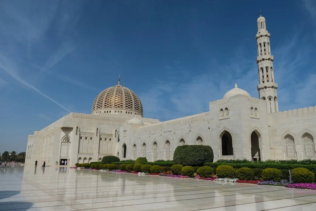 White marble mosque with a large golden dome and smaller domes, surrounded by a courtyard with green bushes and colorful flowers, under a blue sky.