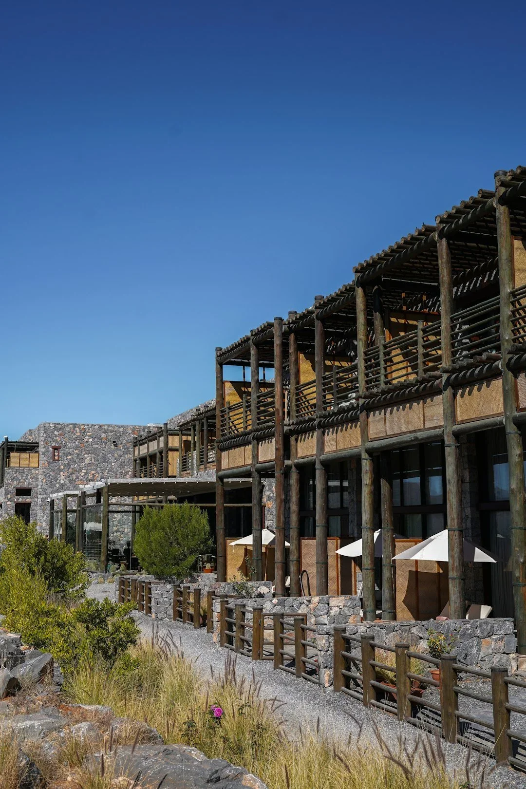 Multi-story building with wooden balconies, stone walls, surrounded by desert plants and rocks, under a clear blue sky.