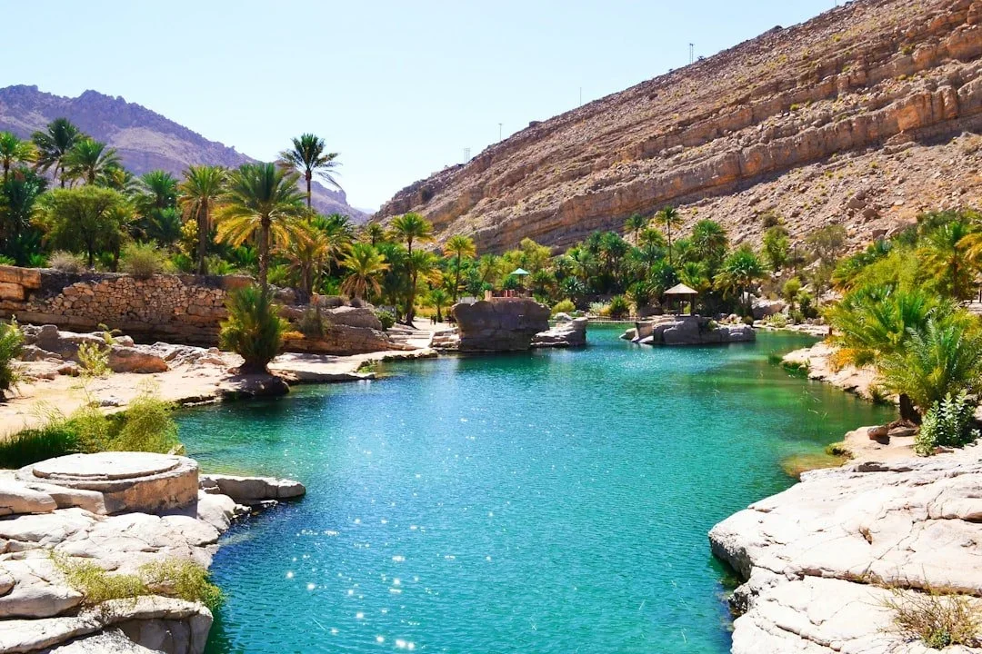 A desert landscape with a turquoise river running through it, surrounded by palm trees and rocky hills in the background under a clear blue sky.