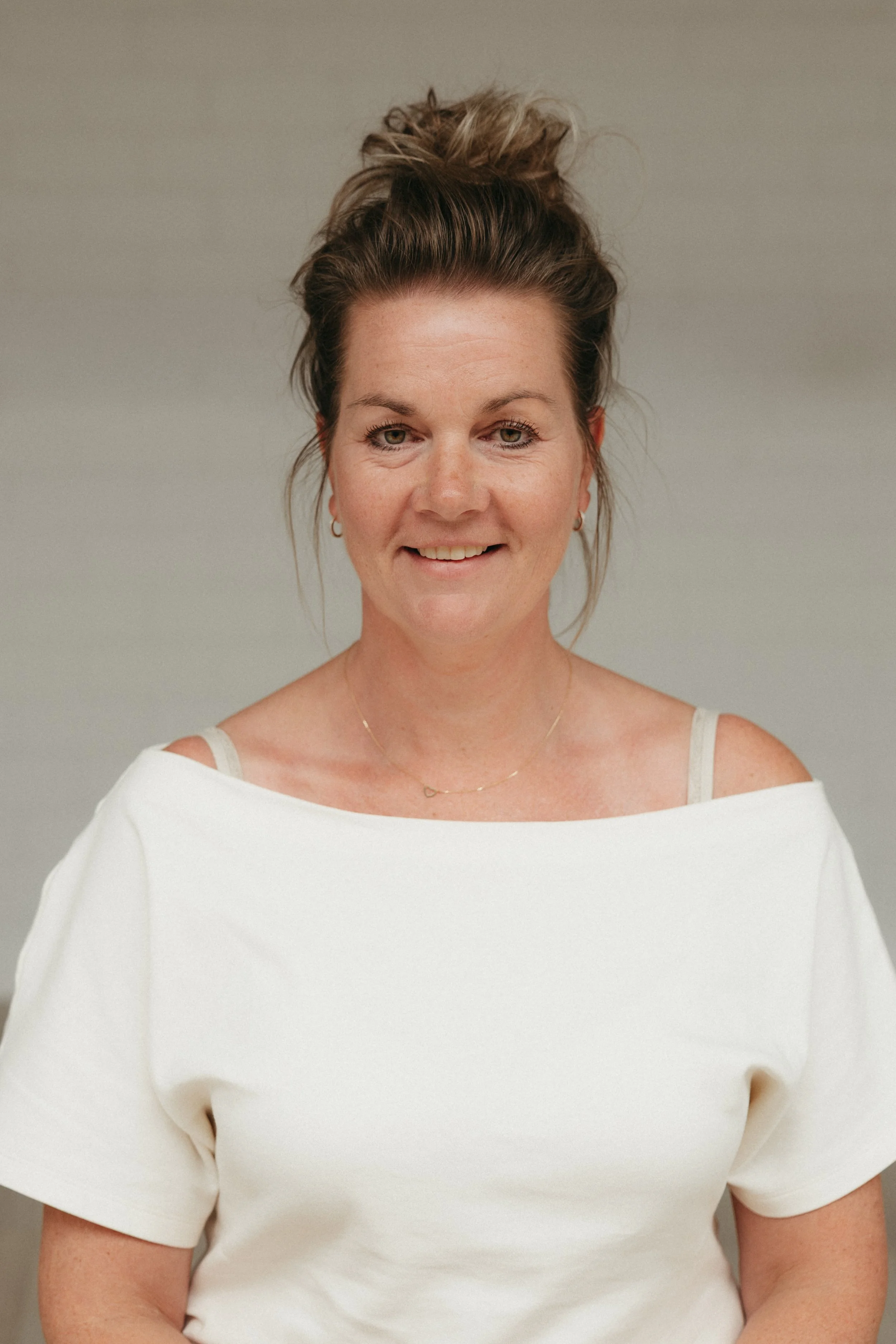 Een vrouw met een losgeschikte, nonchalante paardenstaart en een witte top met schouderbandjes, lachend in een neutrale achtergrond.