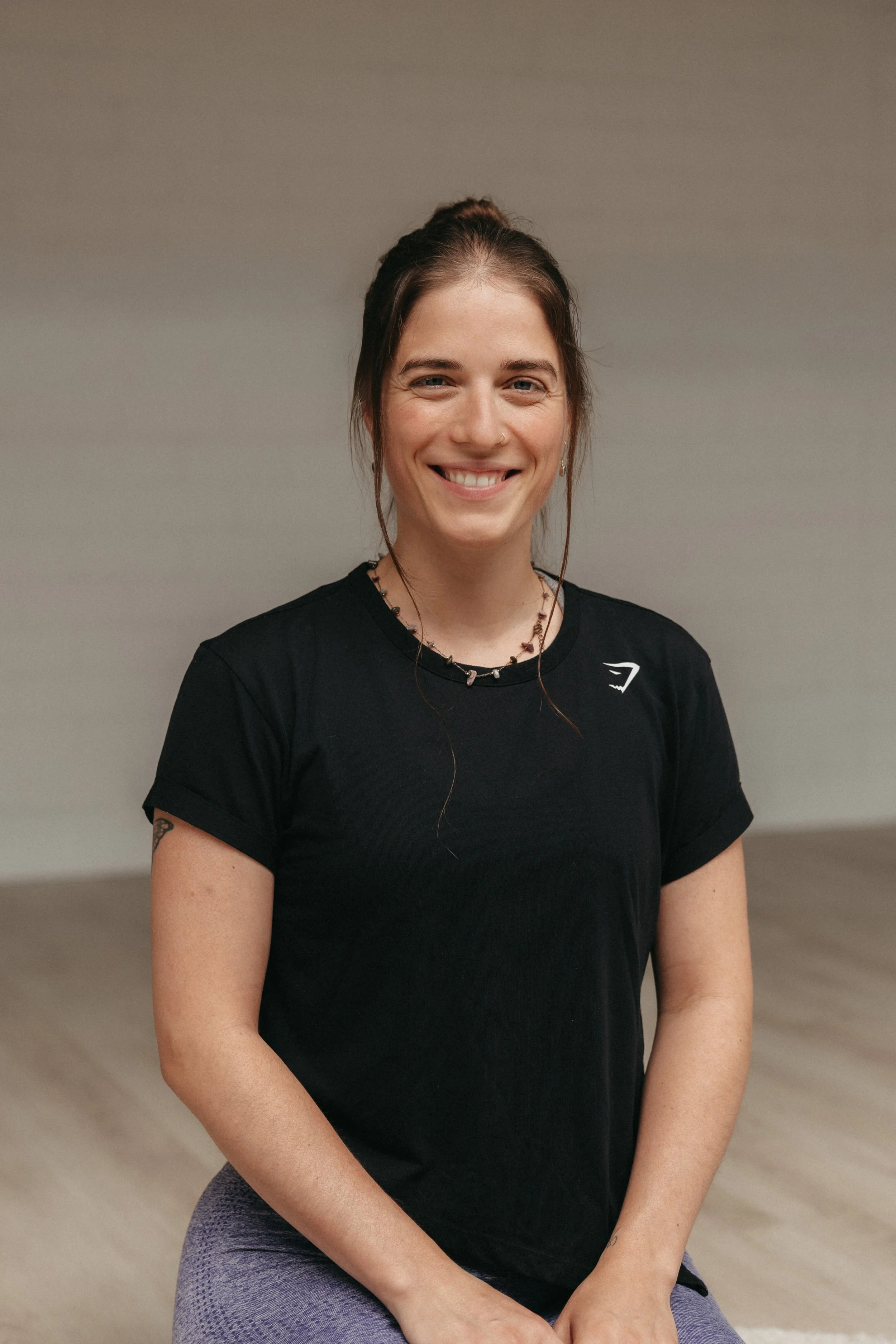 Vrouw met een glimlach, haar haar in een staart, zwarte T-shirt en paarse leggings, zittend op een vloer in een kamer met effen muur.