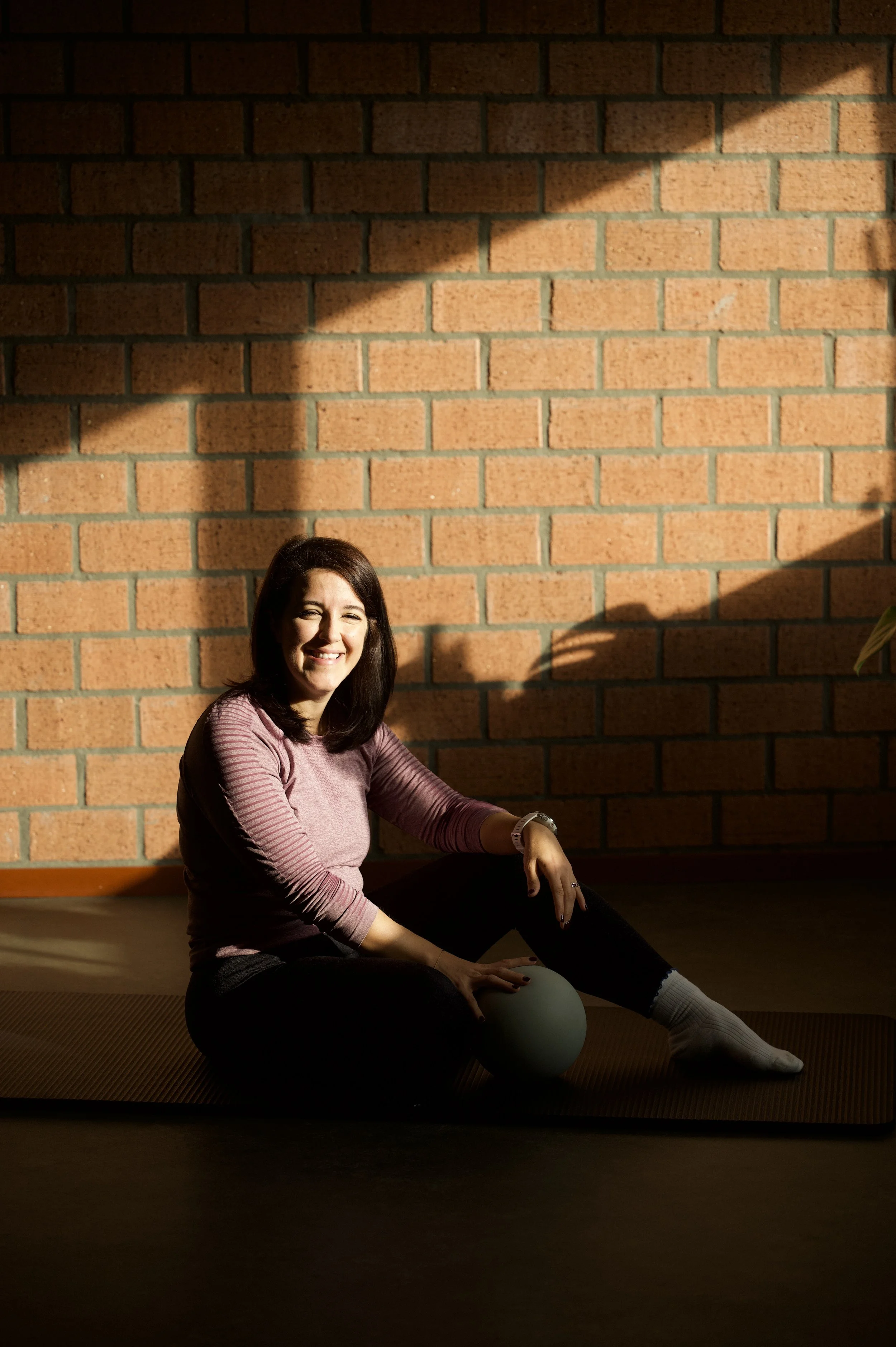 Een lachende vrouw met bruin haar zit op een mat op de vloer, tegen een bakstenen muur, met zonlicht dat over haar heen valt.